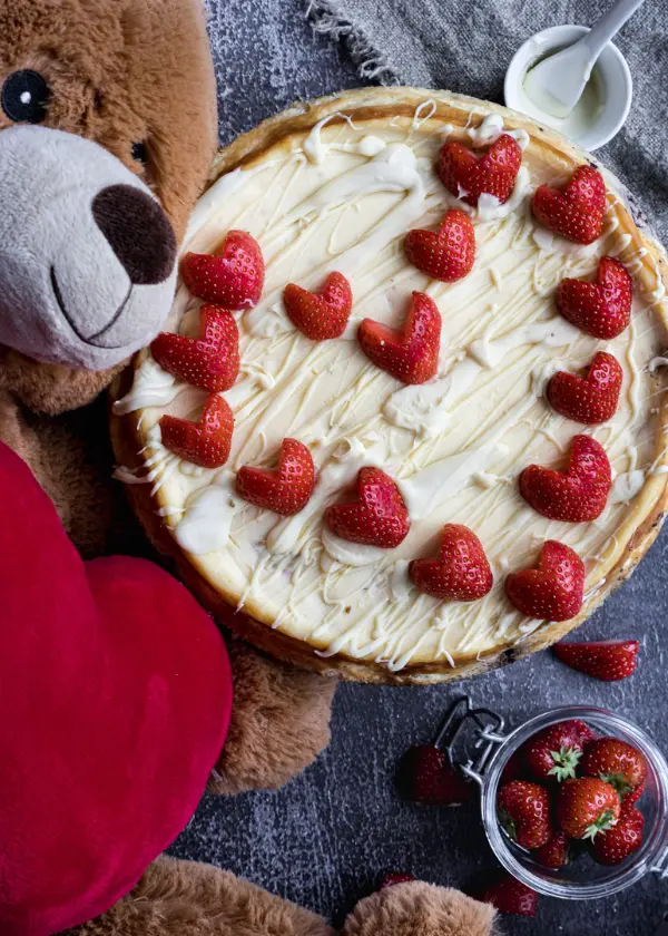Baked white chocolate strawberry cheesecake with heart shaped strawberries on top and a teddy bear on the side